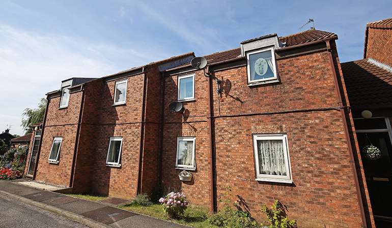 Exterior of a row of two‑storey red‑brick residential homes with white‑framed windows and pitched roofs. Satellite dishes are mounted on the walls, and small front gardens with flowers and shrubs line a paved pavement under a bright blue sky.