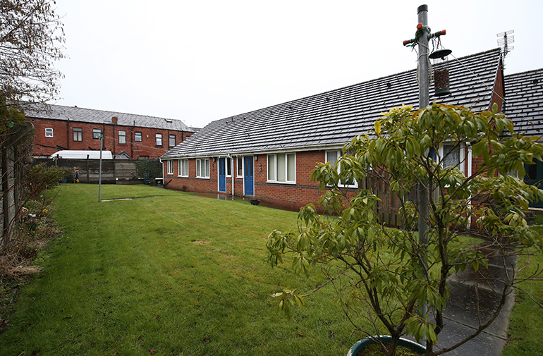 Communal garden beside a single‑storey red‑brick residential building with white‑framed windows and blue front doors. A neatly maintained lawn stretches along the building, with a paved path, a small tree in the foreground, and additional residential buildings visible in the background under an overcast sky.