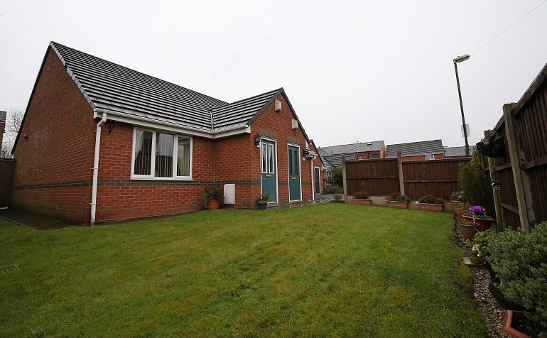 Exterior of a single‑storey red‑brick bungalow with a pitched tiled roof, set within a private fenced garden. A grass lawn fills the foreground, with paved pathways, planters and shrubs arranged around the garden, and neighbouring low‑rise buildings visible beyond the fence.