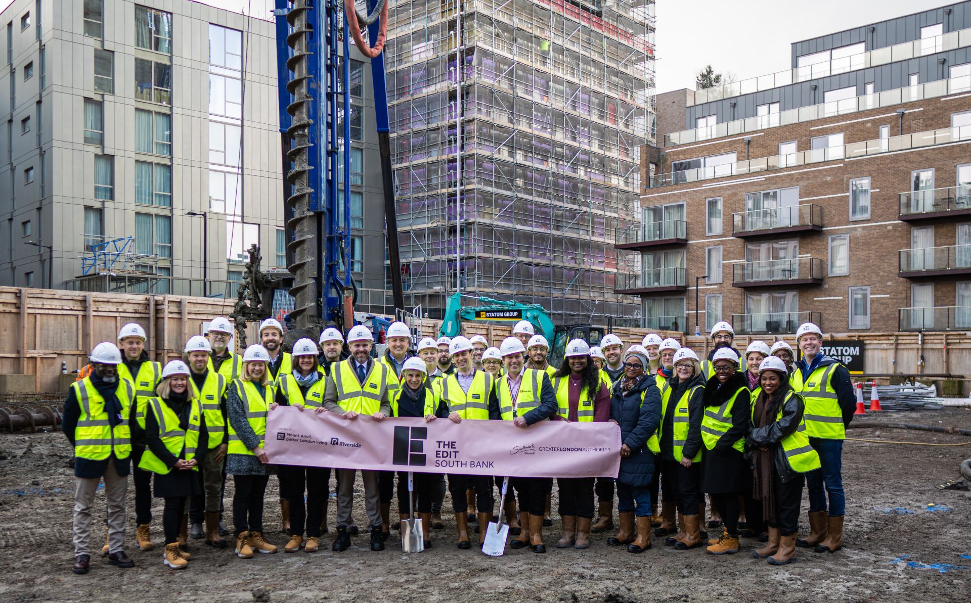 A large group of adults wearing high-visibility vests and hard hats stand on a construction site holding a banner reading “The Edit South Bank.” Behind them are scaffolding, modern apartment buildings, and construction equipment including a drilling rig.