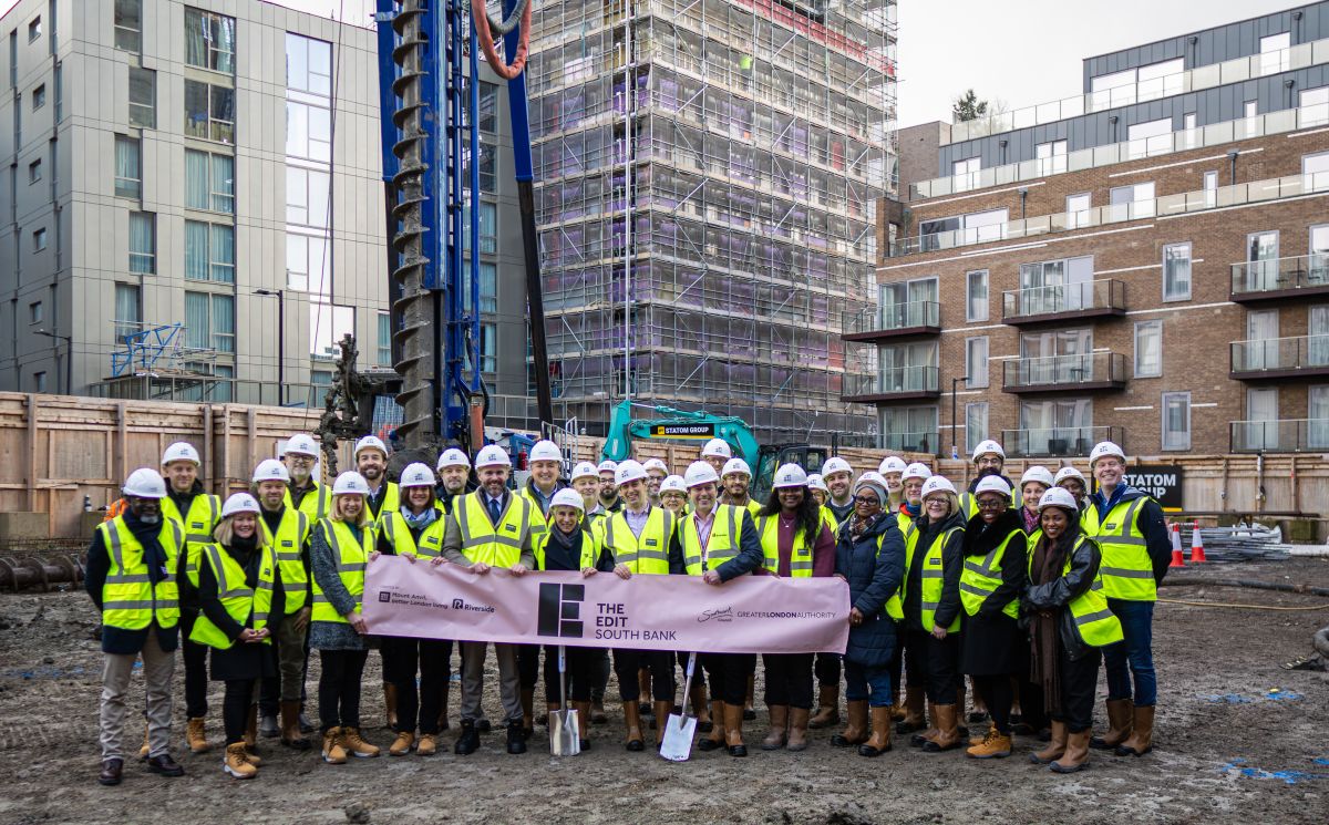 A large group of adults wearing high-visibility vests and hard hats stand on a construction site holding a banner reading “The Edit South Bank.” Behind them are scaffolding, modern apartment buildings, and construction equipment including a drilling rig.