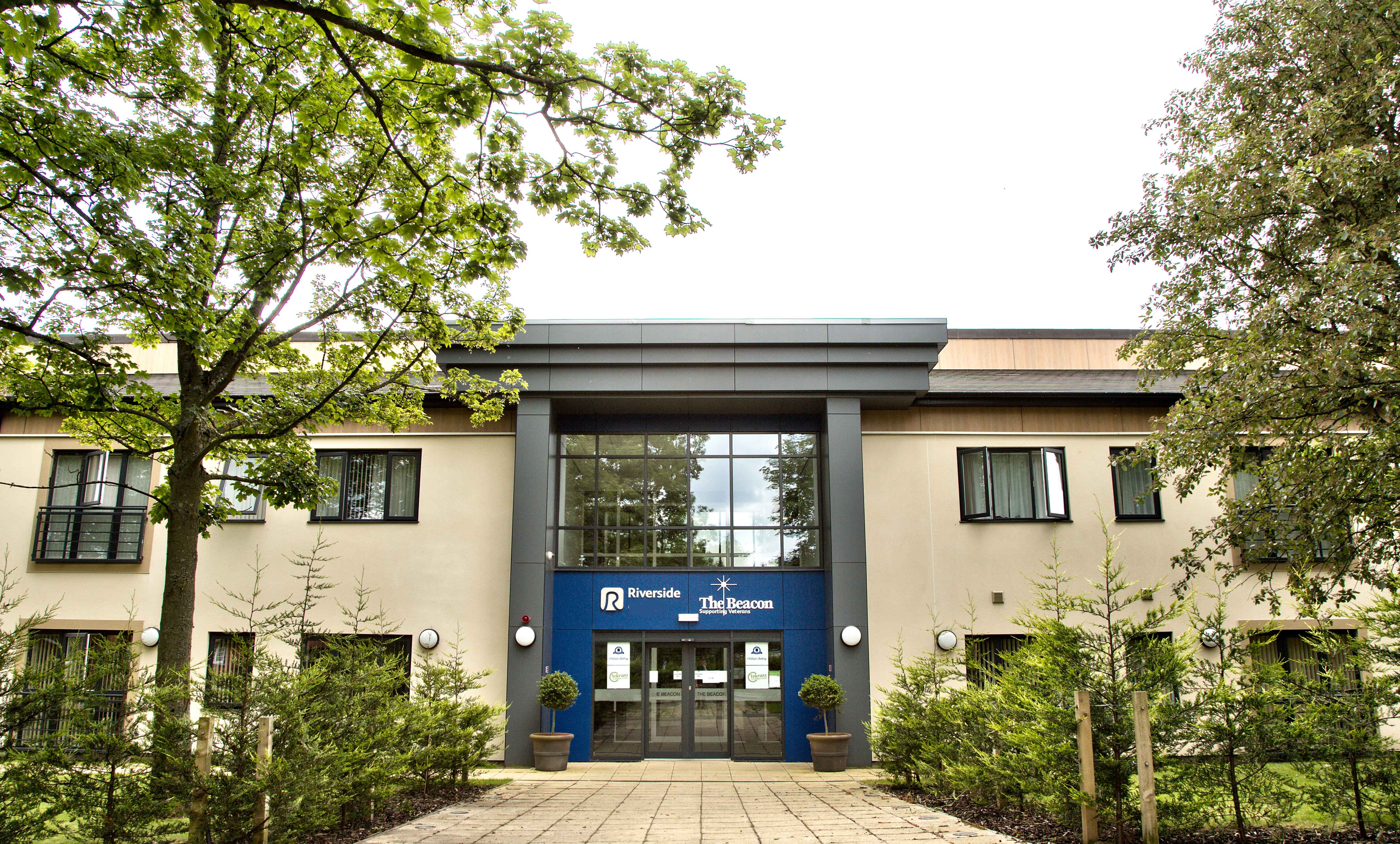 Front entrance of a modern residential building with cream‑coloured walls and a central glass façade. A blue sign above the doors reads “Riverside – The Beacon Supporting Veterans.” A paved pathway leads through landscaped greenery and small trees to the entrance, with upper‑floor windows and balconies visible behind.