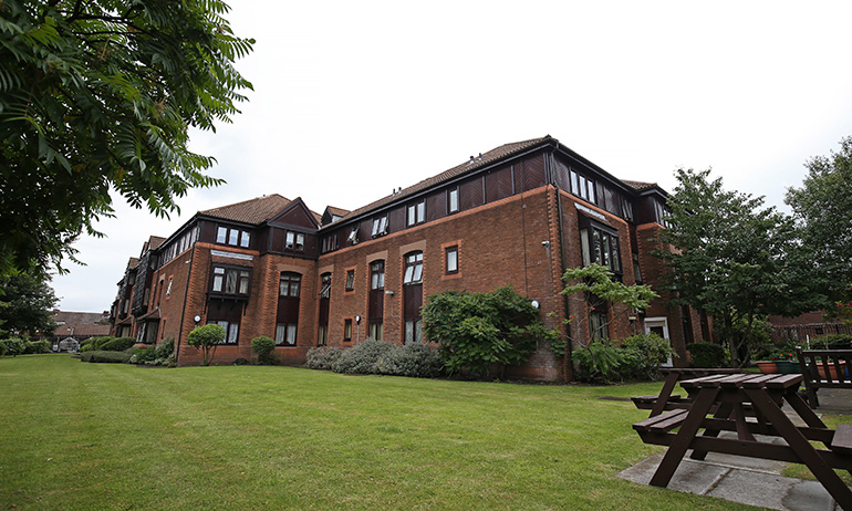 Exterior of a multi‑storey red‑brick residential building with framed windows and dark roof tiles, set beside a large communal lawn. Wooden picnic benches sit on a paved area to one side, with shrubs and trees around the edges and the building overlooking the shared green space under an overcast sky.