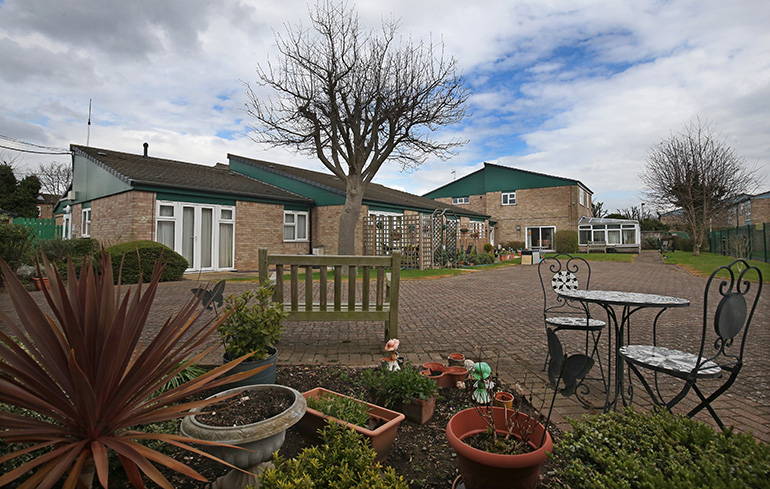 Communal courtyard outside a single‑storey residential building with light‑coloured brick walls and a green roofline. A paved patio area features a wooden bench, metal table and chairs, and multiple potted plants in the foreground, with a trellis, shrubs and additional low‑rise buildings visible around the enclosed garden under an overcast sky.