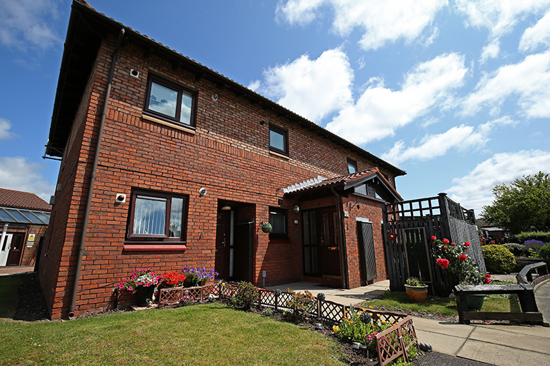 Exterior of a two‑storey red‑brick residential building with wooden‑framed windows and a small covered entrance porch. A neatly maintained front garden features a lawn, flower beds with colourful plants, and low decorative fencing along a paved path leading to the front door, under a bright blue sky with scattered clouds.