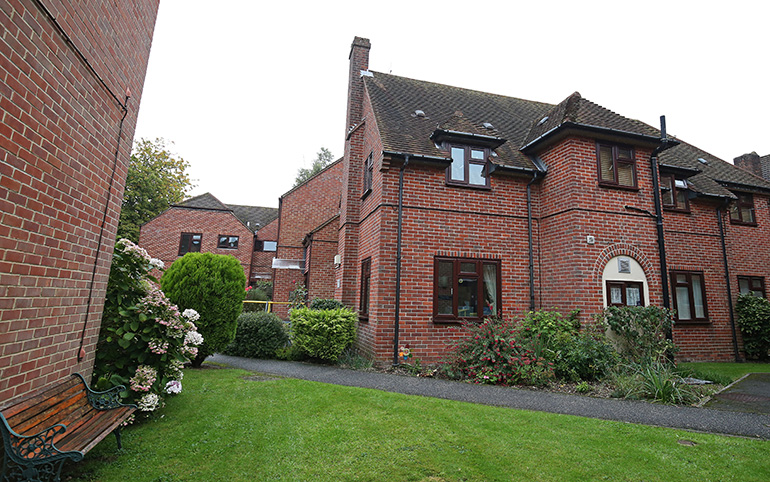 Exterior of a two‑storey red‑brick residential building with white‑framed windows and pitched tiled roofs, set around a landscaped garden. A paved path curves through a neatly maintained lawn, with flowering shrubs and bushes along the building and a wooden bench partially visible in the foreground under an overcast sky.