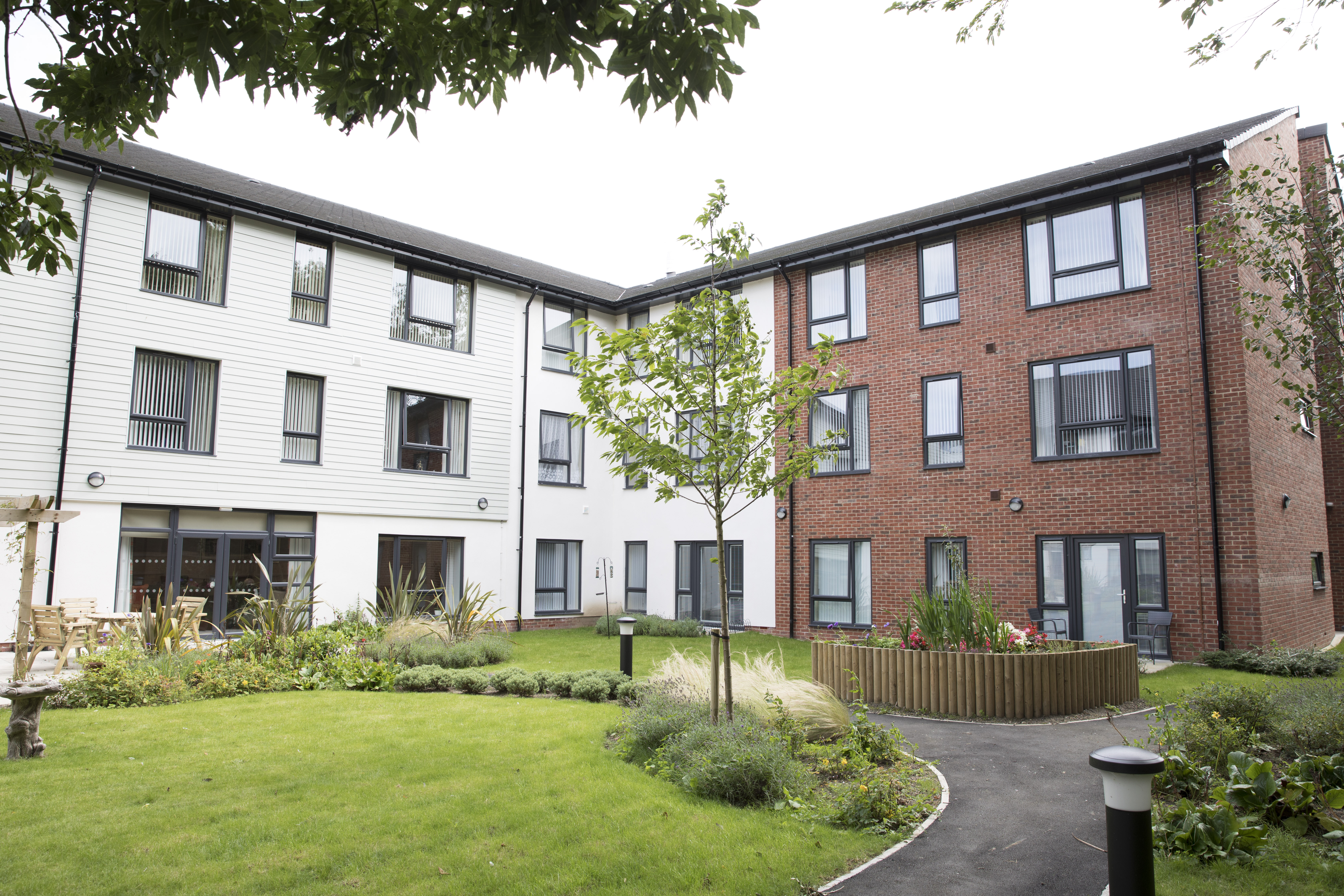 Landscaped courtyard with lawn, trees and raised planting beds, surrounded by modern apartment buildings with white cladding and brick façades.