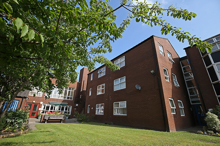 Exterior of a red‑brick multi‑storey residential building viewed across a neatly maintained lawn. A paved path curves through the grass towards a circular feature area with a small central structure. A wooden bench and raised planting beds sit to one side, with windows overlooking the shared outdoor space beneath a bright, lightly clouded sky.
