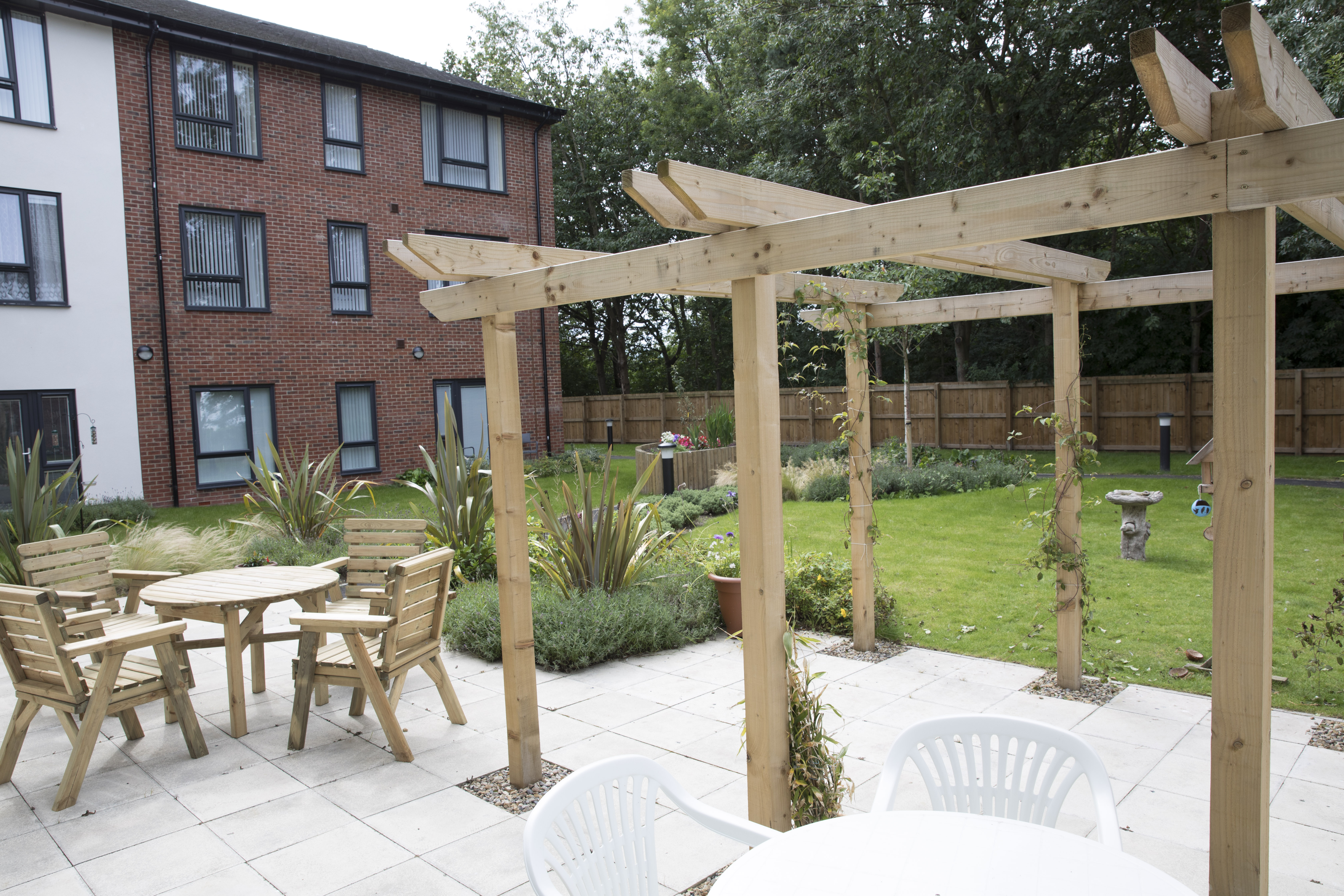 Outdoor seating area with wooden table and chairs beneath a pergola, set within a garden courtyard beside residential buildings.