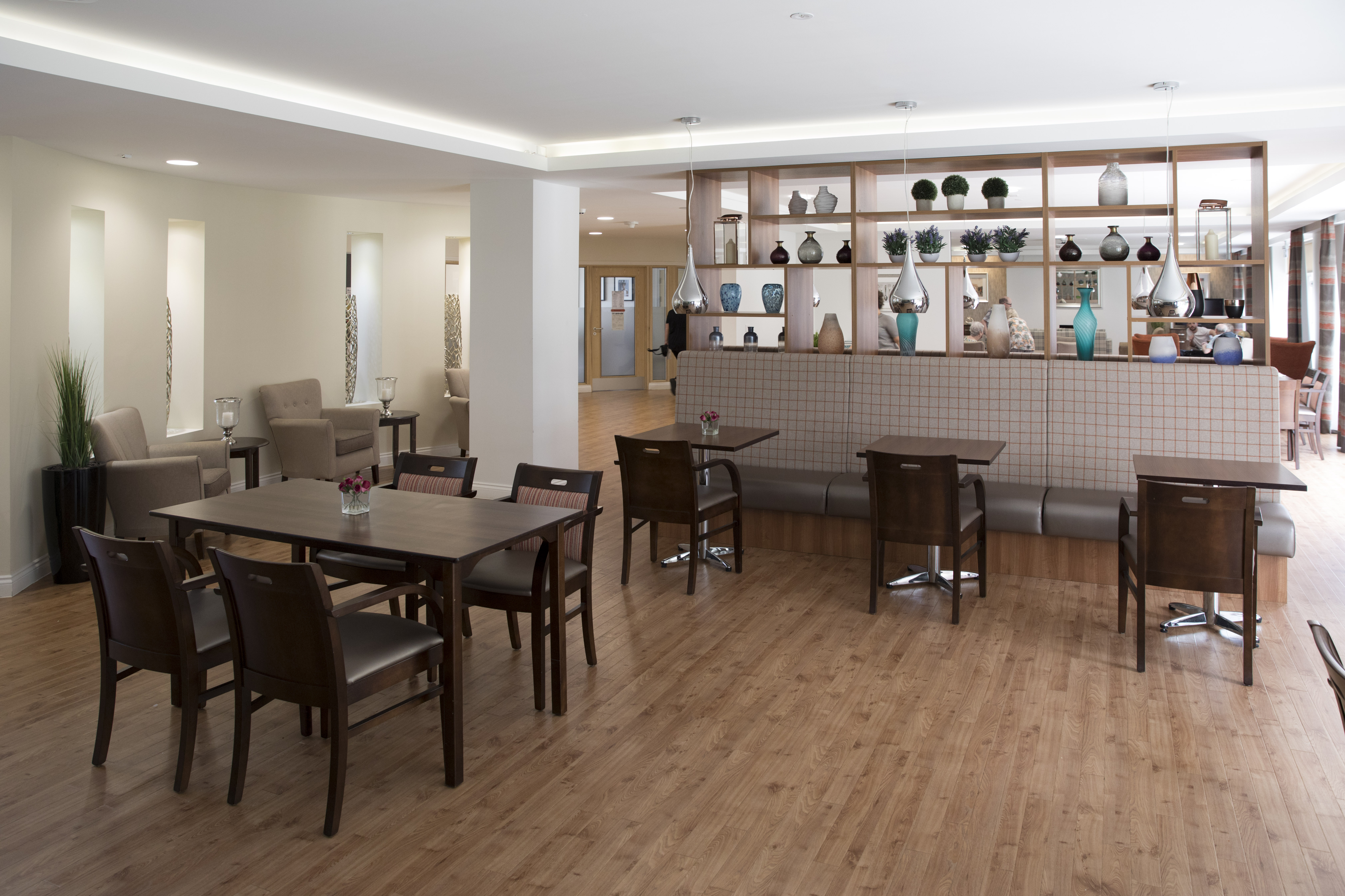 Communal dining and lounge space with tables, chairs and booth seating, separated by shelving with decorative items.