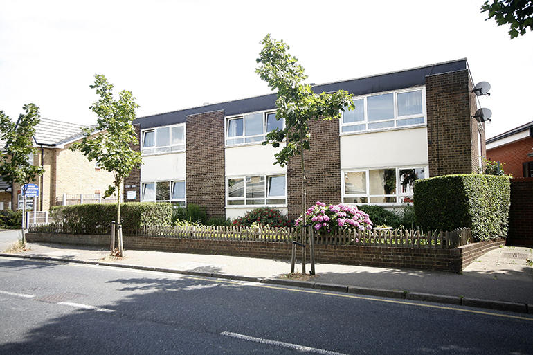 Exterior of a two‑storey residential building with brown brick and cream‑coloured panels, facing a quiet street. A low fence borders a small front garden with flowering shrubs and a young tree, with pavement and road in the foreground.