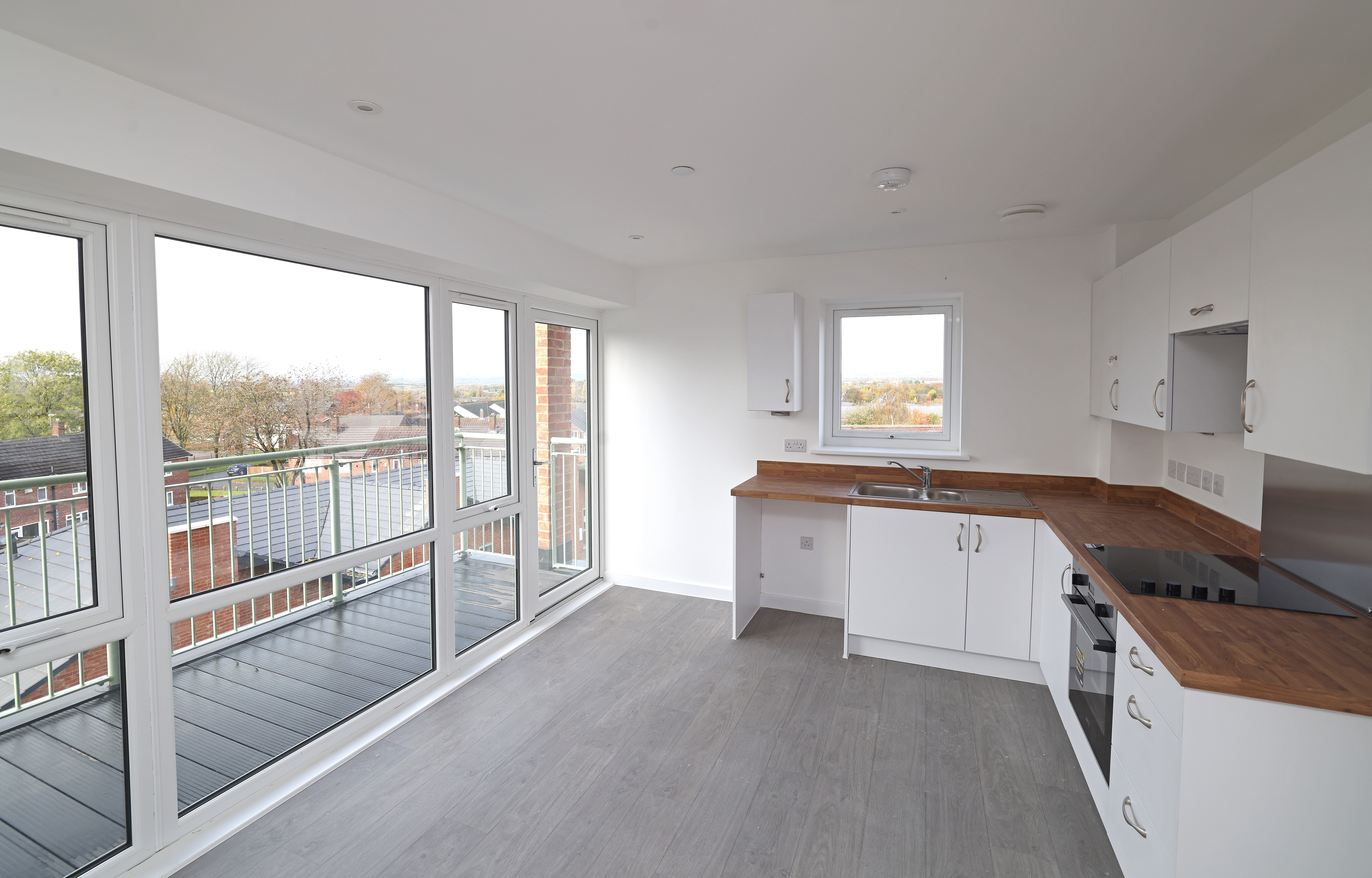 Empty modern kitchen with white units, wooden worktops, integrated oven and large windows leading to a balcony.