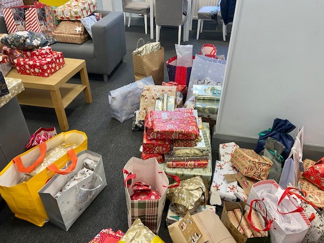 Room filled with stacks of wrapped Christmas presents and gift bags placed on the floor, tables and seating areas. The gifts are wrapped in festive paper and bags, arranged throughout a shared indoor space with chairs, tables and sofas visible.