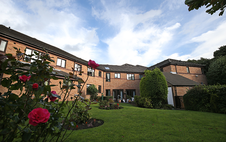 Communal garden in front of a multi‑storey red‑brick residential building, with a wide lawn and curved planting beds featuring blooming rose bushes in the foreground. A paved path leads toward the building entrances, with hedges and a small structure on the right, under a bright sky with scattered clouds.