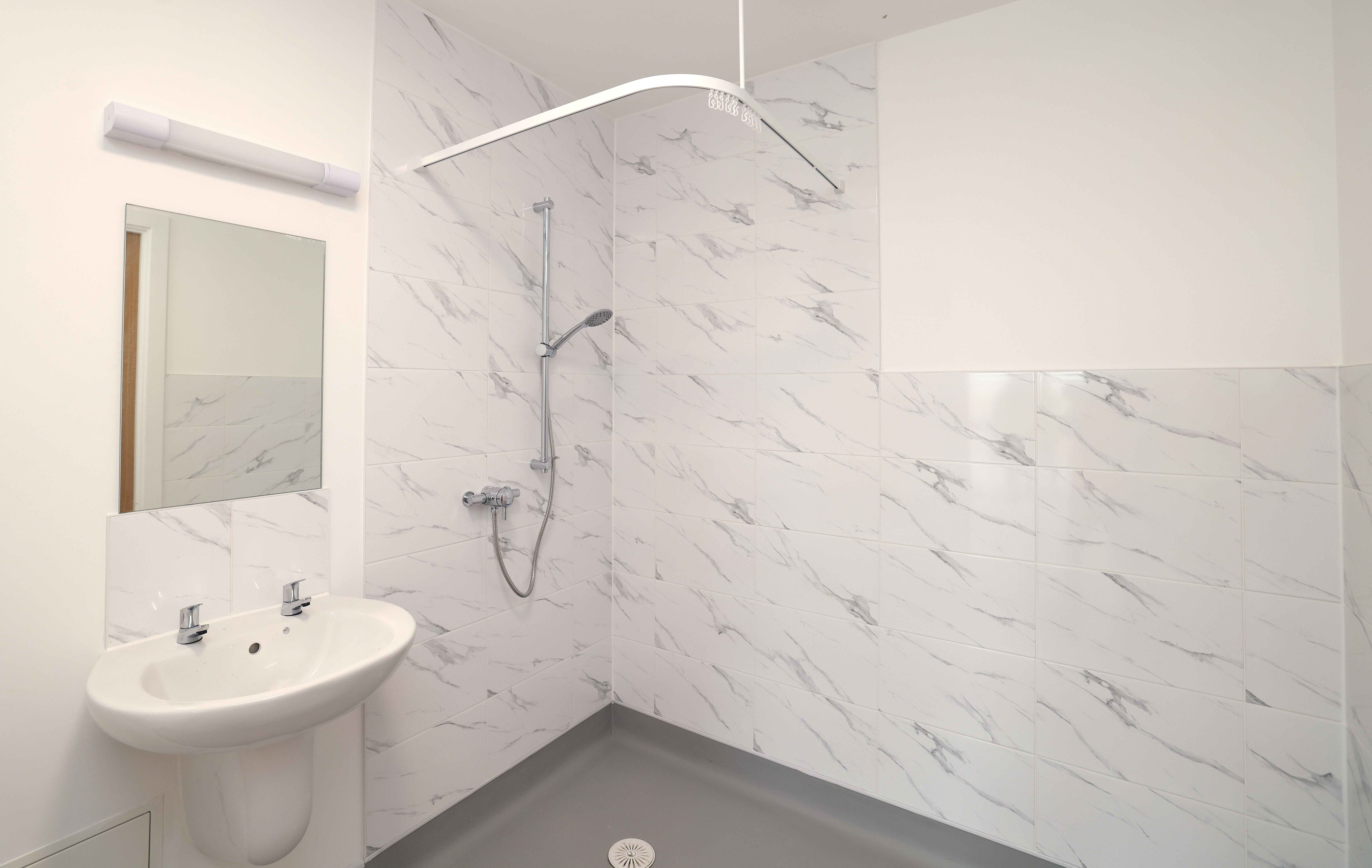 Accessible bathroom with wall-mounted sink, mirror and open walk-in shower with white tiled walls and hand-held showerhead.