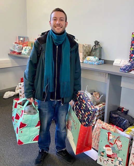 Engage Leeds Toy Appeal Christmas 2022 Nathan Person standing indoors holding two large Christmas gift bags, with wrapped presents and gift bags arranged on tables and the floor behind them. The room has plain walls and shelving, with festive items visible throughout the space.