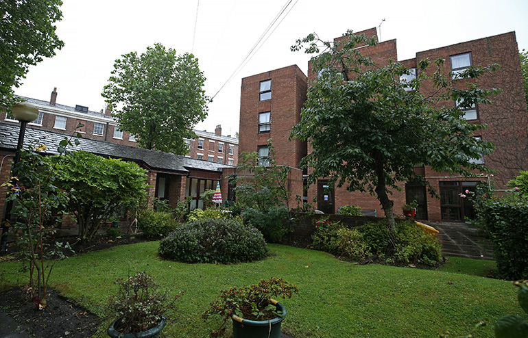Communal garden in front of two red‑brick residential buildings, with a central lawn, shrubs and potted plants. A small paved path leads toward the building entrances, with a mature tree and additional planting throughout the shared outdoor space under an overcast sky.