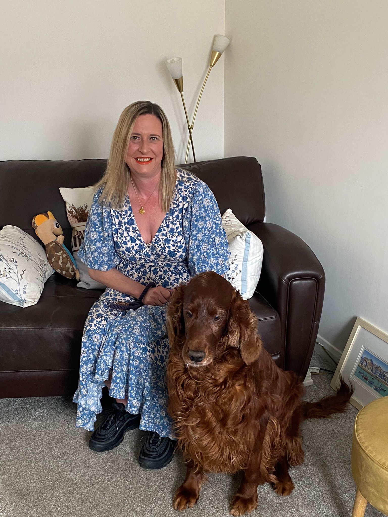 Person seated on a sofa in a living room wearing a blue patterned dress, with a brown dog sitting in front of them. Cushions, a floor lamp and framed picture are visible in the background.