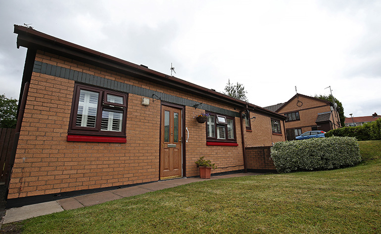 Exterior of a single‑storey brick bungalow with a shallow pitched roof, set beside a neatly mown lawn. The property has white‑framed windows, a wooden front door with a small hanging basket, and low hedging nearby, with another bungalow visible in the background under a cloudy sky.