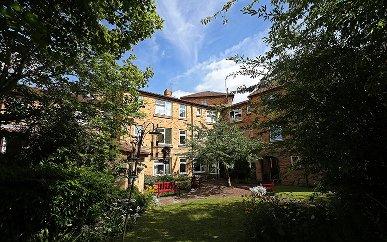 Enclosed garden courtyard surrounded by red‑brick residential buildings with white‑framed windows. A red bench sits among lawns, flower beds and shrubs, with trees and leafy planting framing the space beneath a blue sky with light clouds.