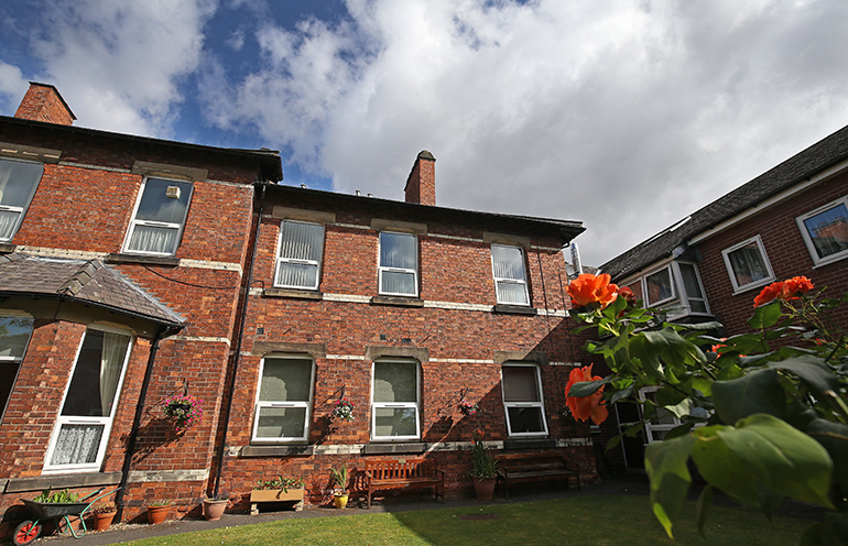 Exterior of a red‑brick residential building with white‑framed windows, viewed across a small communal garden. Wooden benches, potted plants and hanging baskets line the building, with a neatly kept lawn in the foreground, orange flowers close to the camera, and a partly cloudy sky overhead.