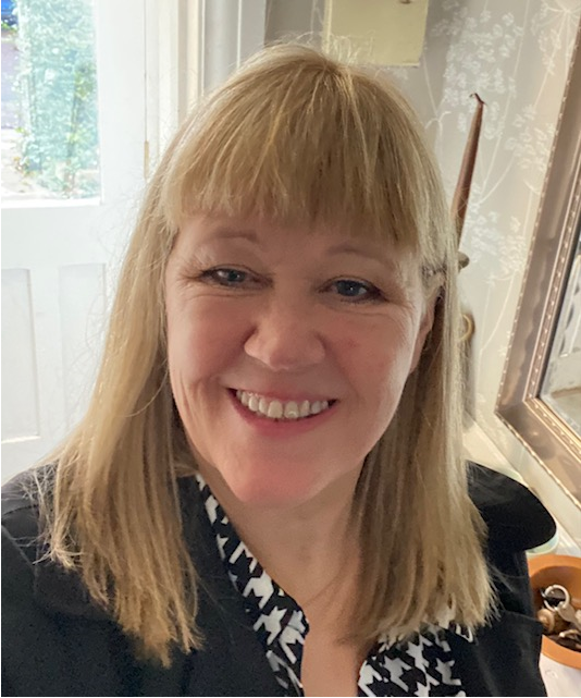 Portrait of a person with shoulder‑length light hair and fringe, wearing a dark jacket over a patterned top. The photo is taken indoors, with a doorway and mirror visible in the background.