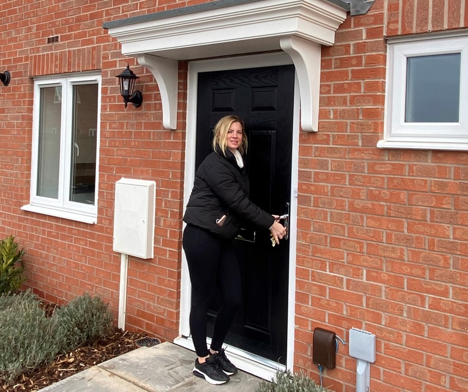 Person standing at the front door of a red‑brick house, opening a black front door beneath a white porch canopy. A paved path, small shrubs and ground‑floor windows are visible on the exterior of the home.