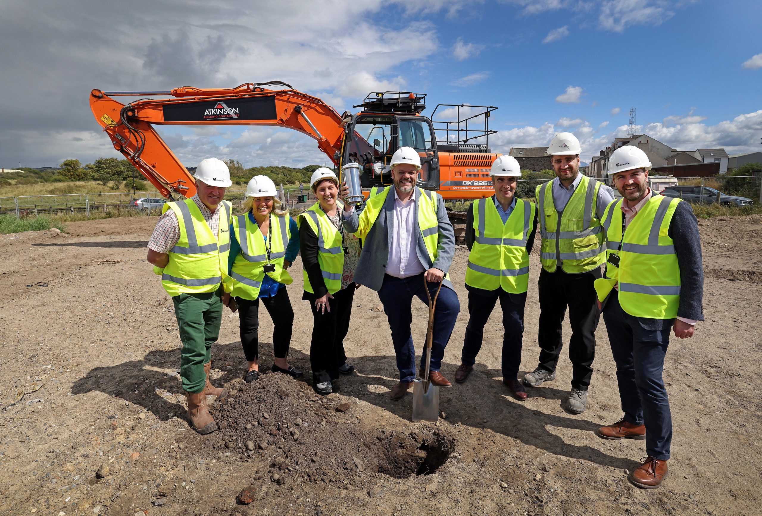 Group of people wearing high‑visibility jackets and safety helmets stand together at a ground‑breaking event on a construction site. One person holds a spade and a time capsule above freshly turned soil, with an excavator and open land visible behind them under a partly cloudy sky.