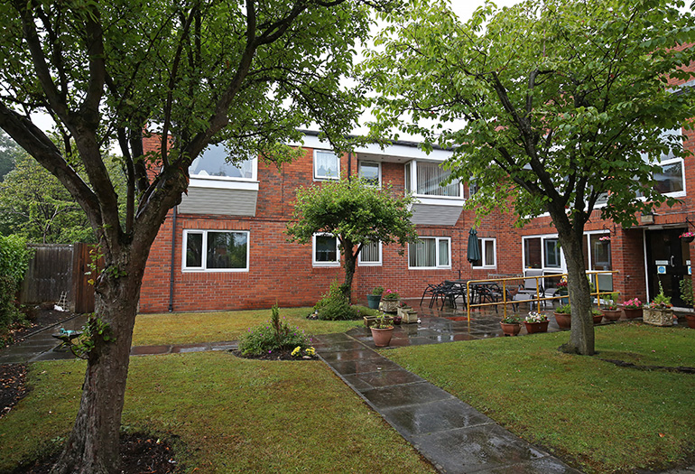 Communal garden outside a red‑brick residential building, with a paved pathway leading through a lawn toward outdoor seating and planters. Mature trees frame the space, and ground‑floor and upper‑floor windows overlook the shared garden area.