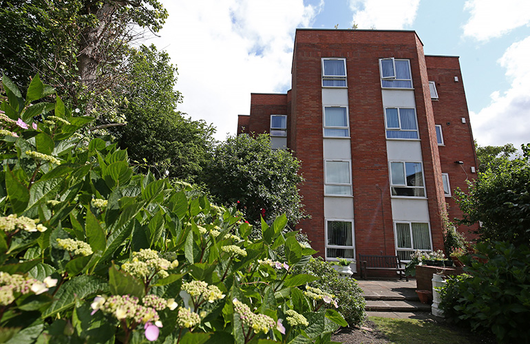 Multi‑storey red‑brick residential building viewed from a landscaped garden. Flowering shrubs and leafy plants fill the foreground, with a paved path and bench near the base of the building and tall white‑framed windows rising above the greenery under a bright sky.