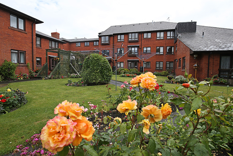 Landscaped communal garden in front of red‑brick residential buildings, with orange and yellow roses in the foreground. A central lawn, flower beds and a rotary clothes line sit within the shared green space, with windows overlooking the garden from surrounding buildings.