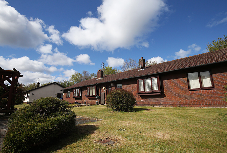 Row of single‑storey red‑brick bungalows with pitched tiled roofs and white‑framed windows, set behind a gently sloping lawn. Low shrubs and small bushes are planted across the grass, with chimneys along the roofline and trees and clouds visible against a blue sky.