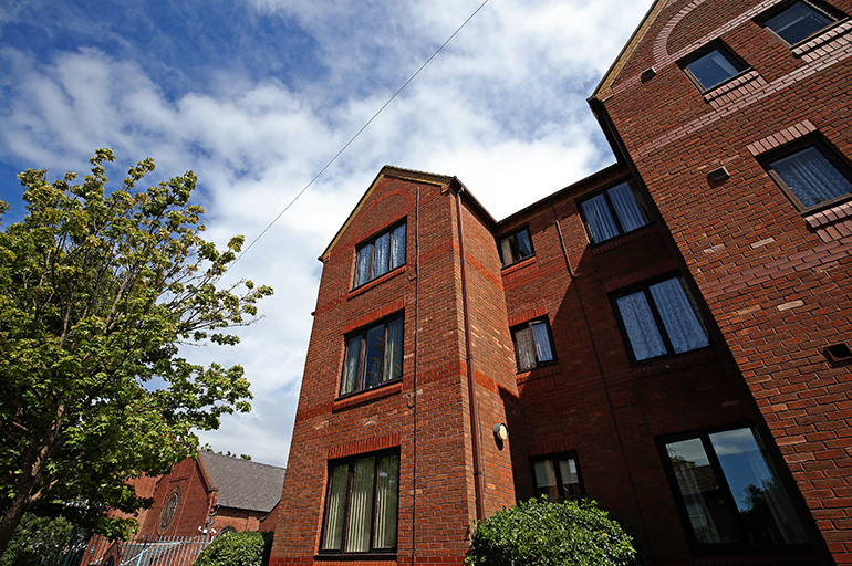 Exterior view of a multi‑storey red‑brick residential building with white‑framed windows, photographed from below against a partly cloudy blue sky. A mature leafy tree stands beside the building, with shrubs at ground level and neighbouring buildings visible in the background.