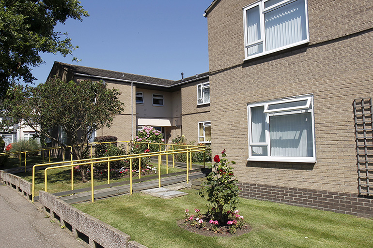 Exterior of a two‑storey residential building with light brown brick walls and white‑framed windows. A gently sloped access ramp with yellow handrails leads to the entrance across a neatly cut lawn, with small flower beds, shrubs and a low concrete wall along the pathway, under a clear blue sky.