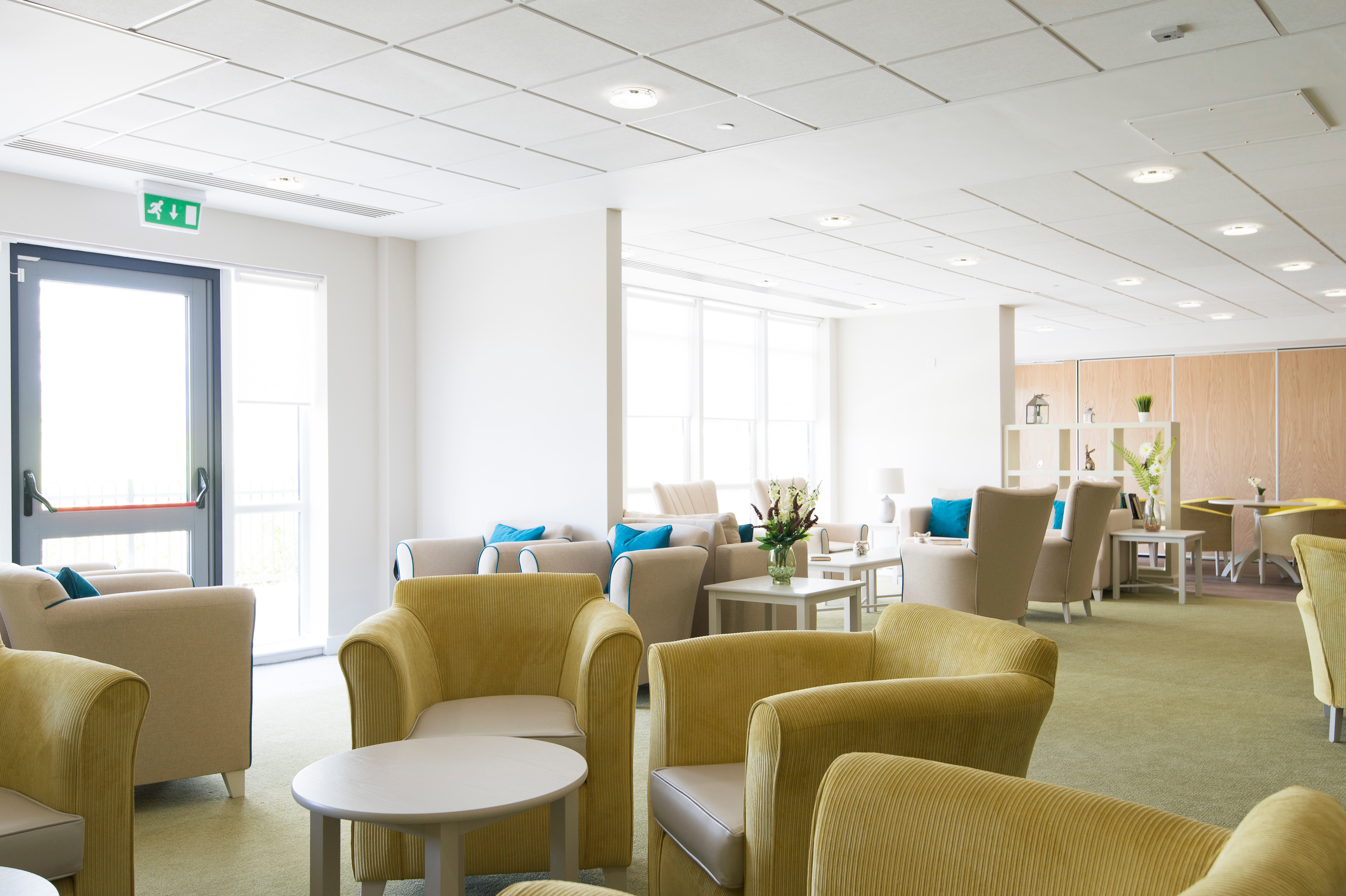 Bright communal lounge with armchairs, small tables and large windows, arranged as a shared seating area inside a care facility.