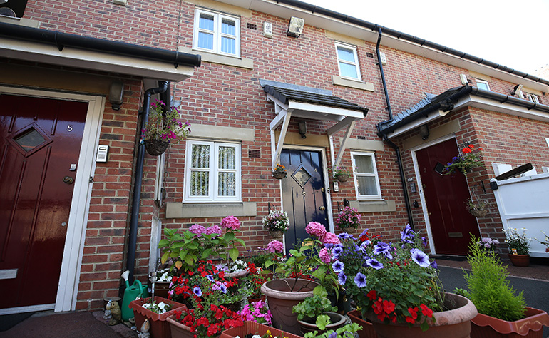 Exterior of two‑storey red‑brick terraced houses with painted front doors and small porches. Colourful potted flowers and hanging baskets are arranged outside the entrances, creating a well‑maintained residential frontage.