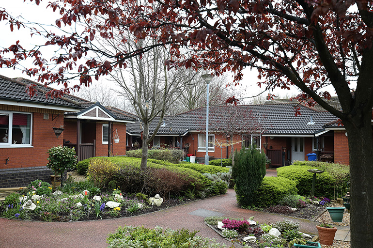 Communal garden serving a group of single‑storey red‑brick bungalows, with curving paved pathways running through landscaped flower beds and shrubs. Small trees and a bird table sit within the shared green space, with front doors and windows facing onto the garden area.