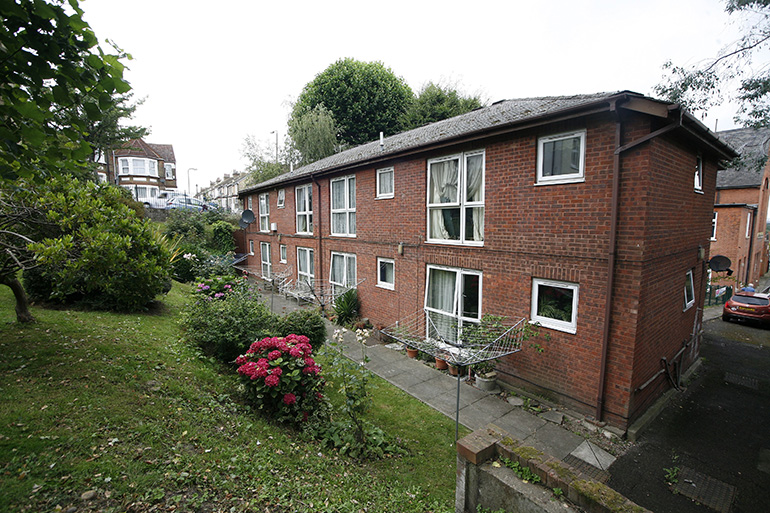Exterior of a two‑storey red‑brick residential building with white‑framed windows, viewed along a gently sloping paved path. A small communal garden with shrubs and flowering plants runs alongside the building, with lawns and mature greenery around the site and neighbouring houses visible in the background.