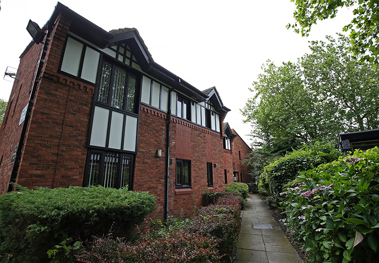 Exterior of a red‑brick residential building with distinctive black‑and‑white timber‑style detailing on the upper windows. A narrow paved path runs alongside the building through landscaped planting beds with shrubs and flowers, bordered by dense greenery and trees, with the building extending into the background under a bright sky.