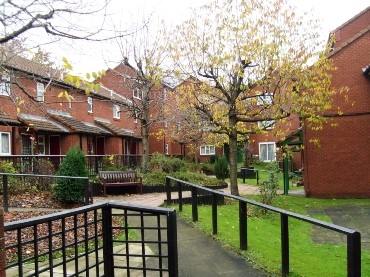 Residential courtyard with a paved pathway and black metal handrails running through a landscaped green space. Red‑brick houses with white‑framed windows face the garden, which includes benches, trimmed lawns, shrubs and a small tree with autumn leaves under an overcast sky.