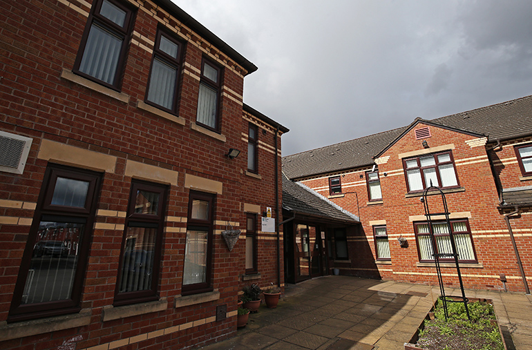 Exterior courtyard of a red‑brick residential building with white‑framed windows and decorative brick detailing. A paved walkway leads between connected buildings toward a covered entrance, with potted plants, small planting beds and low walls around the shared outdoor space under an overcast sky.
