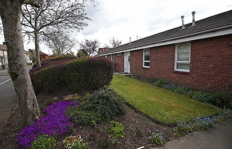 Exterior of a single‑storey red‑brick bungalow with white‑framed windows and a pitched tiled roof. A neatly maintained front garden includes a grass lawn, low hedges and colourful flowering plants, with a paved path leading to the front door and mature trees lining the pavement under an overcast sky.