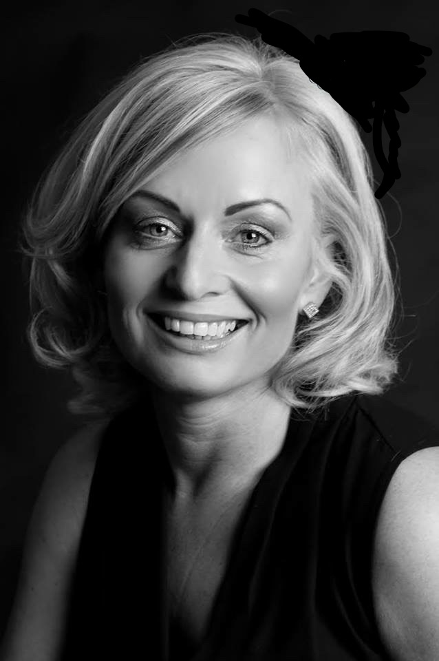 Black‑and‑white studio portrait of a person with short, light‑coloured hair, wearing a sleeveless dark top. They face the camera against a dark background, with even lighting highlighting their face and shoulders.