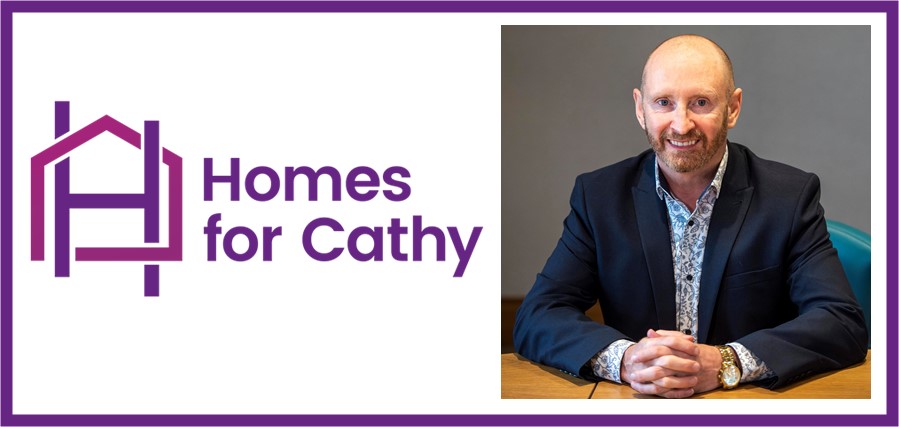 Graphic featuring the Homes for Cathy logo on the left in purple, with a house‑shaped icon and the text “Homes for Cathy.” On the right is a portrait of a person seated at a table indoors, wearing a dark jacket and patterned shirt, with hands resting on the tabletop against a plain background.