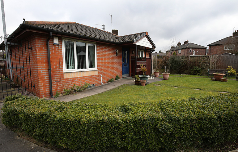 Exterior of a single‑storey red‑brick bungalow with a pitched tiled roof, set behind a neatly maintained lawn. A paved path leads to the front door, with potted plants near the entrance and hedging along the boundary, and neighbouring homes visible in the background.