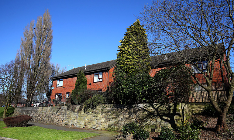 Exterior of a red‑brick residential building set above a stone retaining wall, surrounded by mature trees and shrubs. A grassy area and footpath run along the front of the property, with balconies and windows overlooking the landscaped surroundings under a clear blue sky.