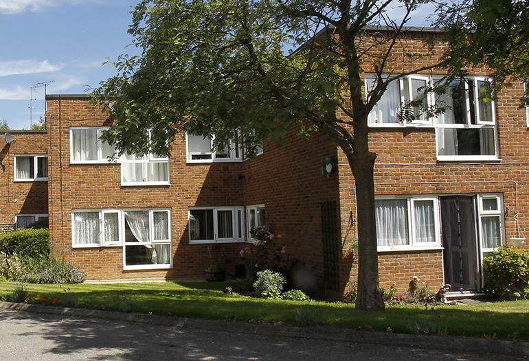 Exterior of a two‑storey red‑brick residential building with white‑framed windows, set within a landscaped garden. A mature tree stands in the foreground beside a neatly kept lawn, with shrubs and flowering plants around the base of the building under a bright sky.