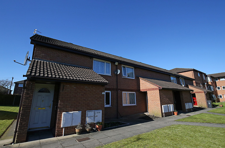 Exterior of a two‑storey red‑brick residential building with pitched tiled roofs and covered entrance porches. A paved path runs along the front, bordered by a neatly cut grass lawn, with adjacent similar buildings visible under a clear blue sky.
