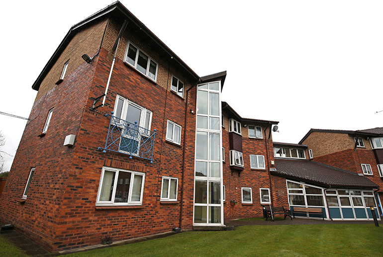Exterior of a multi‑storey red‑brick residential building with white‑framed windows and a vertical glass stairwell feature. A small grass lawn sits in the foreground, with benches and pathways near the entrance and neighbouring buildings visible around the site under an overcast sky.
