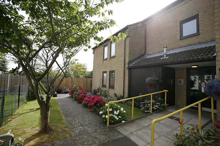 Exterior of a two‑storey residential building with light‑coloured brick walls and a covered entrance. A paved pathway and ramp with yellow handrails lead to the doorway, bordered by neatly planted flower beds with pink and white flowers, a small tree, and a fenced garden area beside the building.